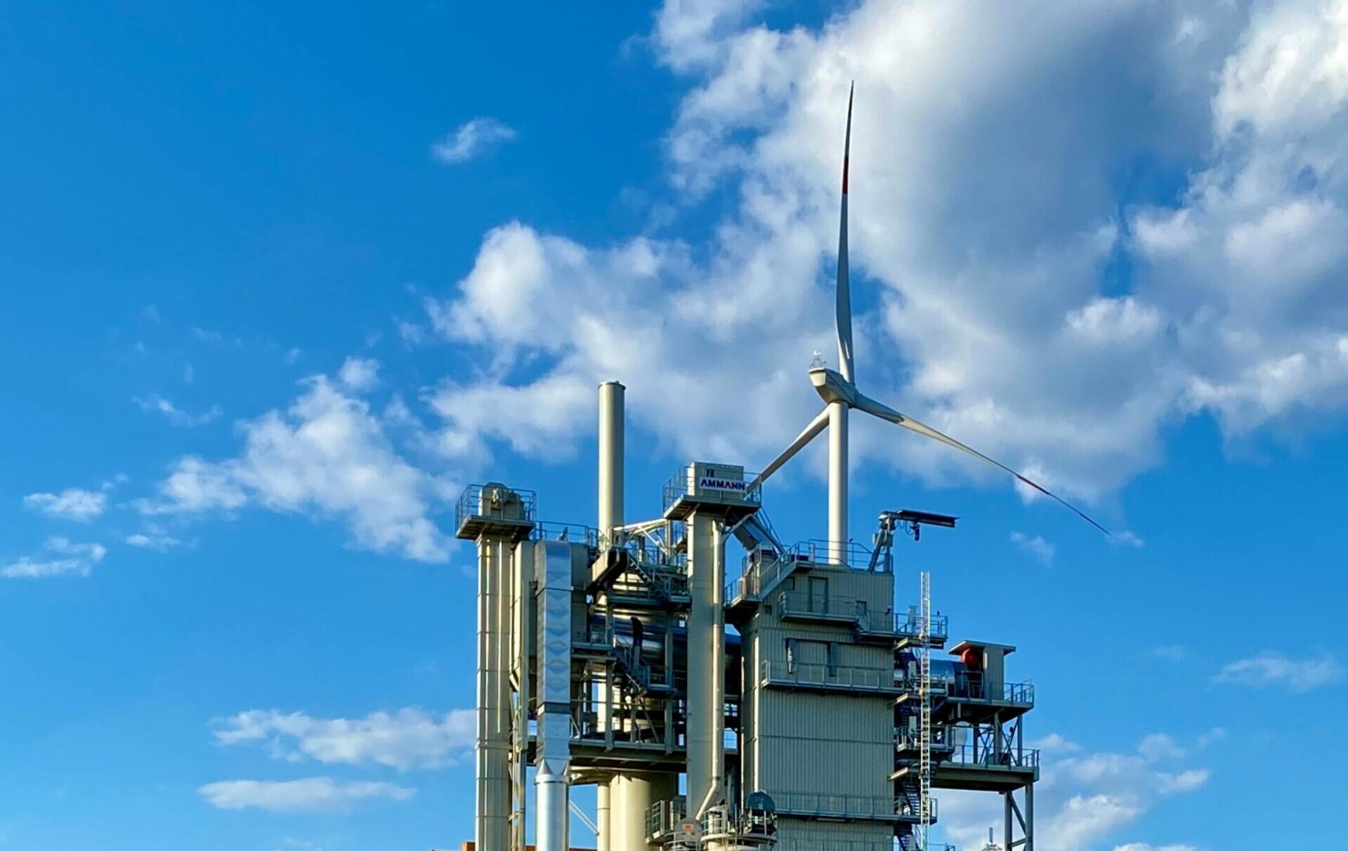Industrial plant with wind turbine, solar panels, and graffiti by a river under a bright blue sky.